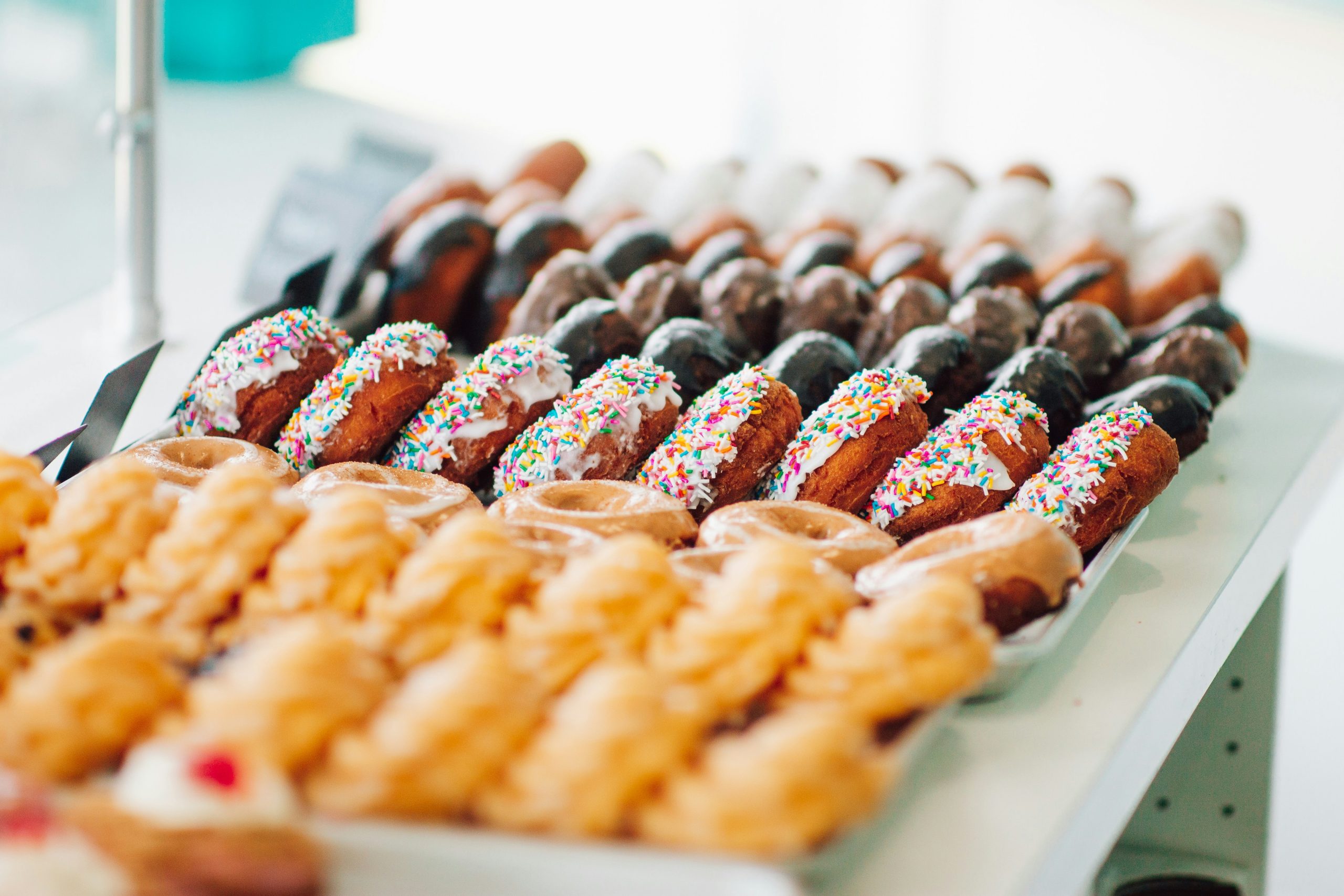 7 rows of assorted donuts—including glazed, powdered, chocolate, and sprinkled—displayed in front of a sunlit window on a wooden table.