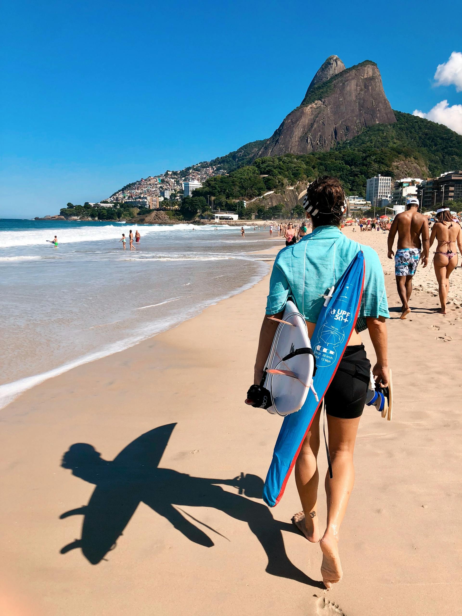 A surfer carrying a board strolls down Ipanema Beach, Rio de Janeiro, under a sunny sky, with a scenic mountain backdrop.