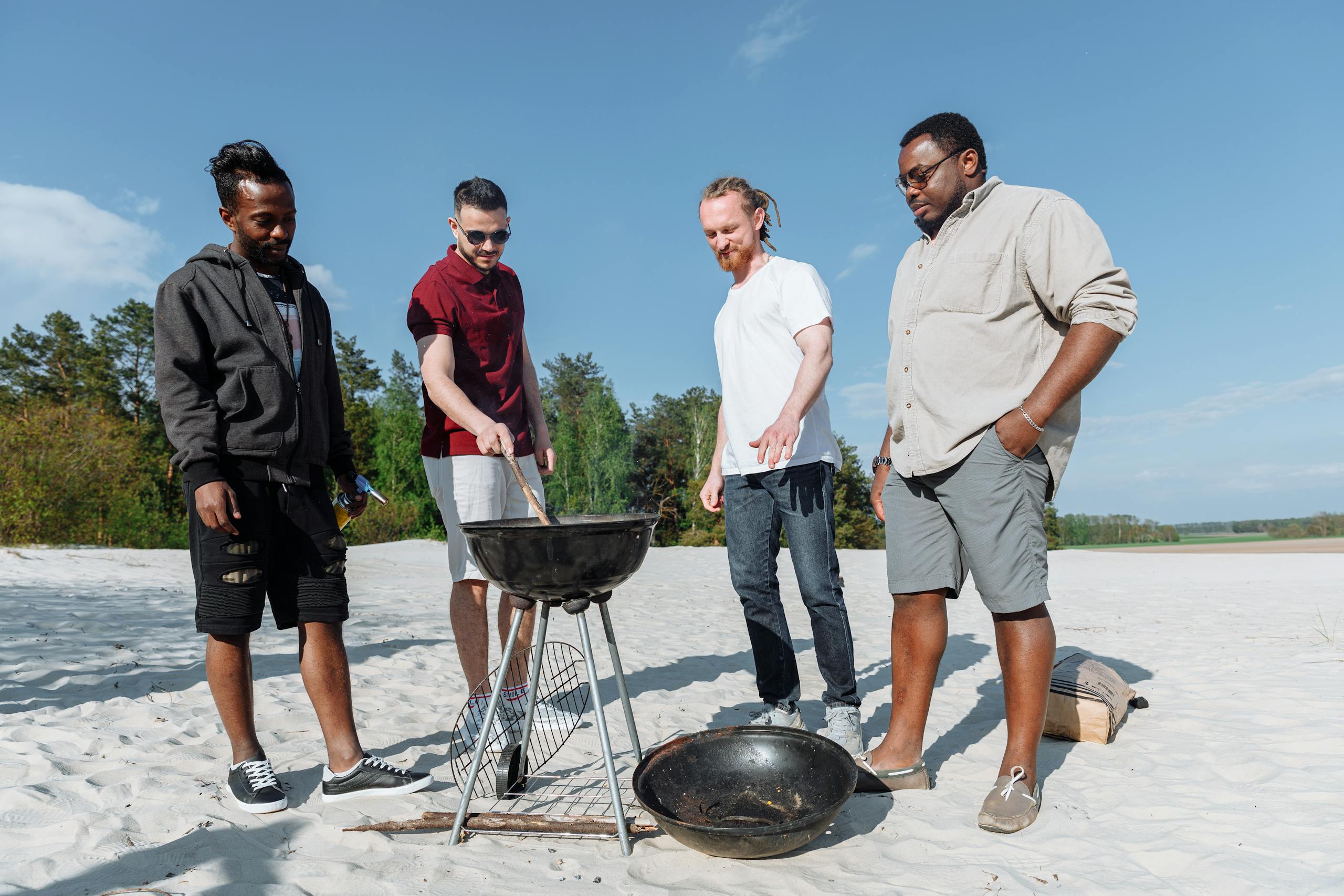 Group of diverse friends grilling together on a sandy beach under a clear blue sky.